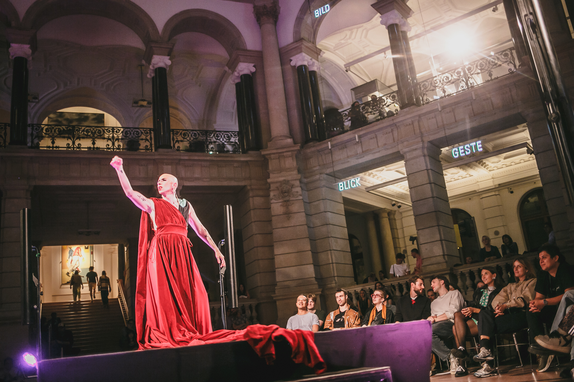 A performer in red attire performs on stage before an audience in an elegant room.