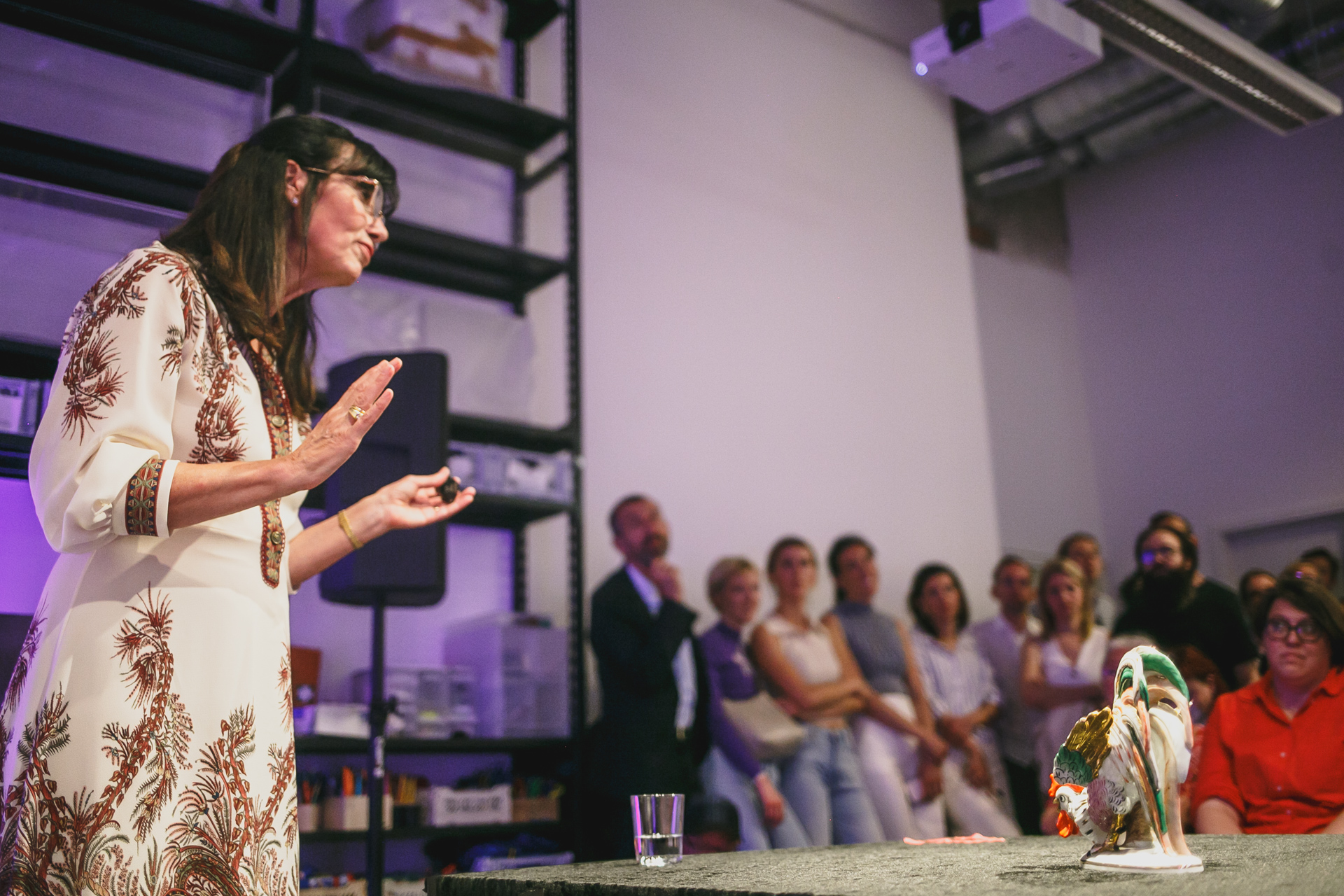 A woman (Heide Rezepa Zabel) speaks to an audience while presenting an artistic figure. Listeners are visible in the background.