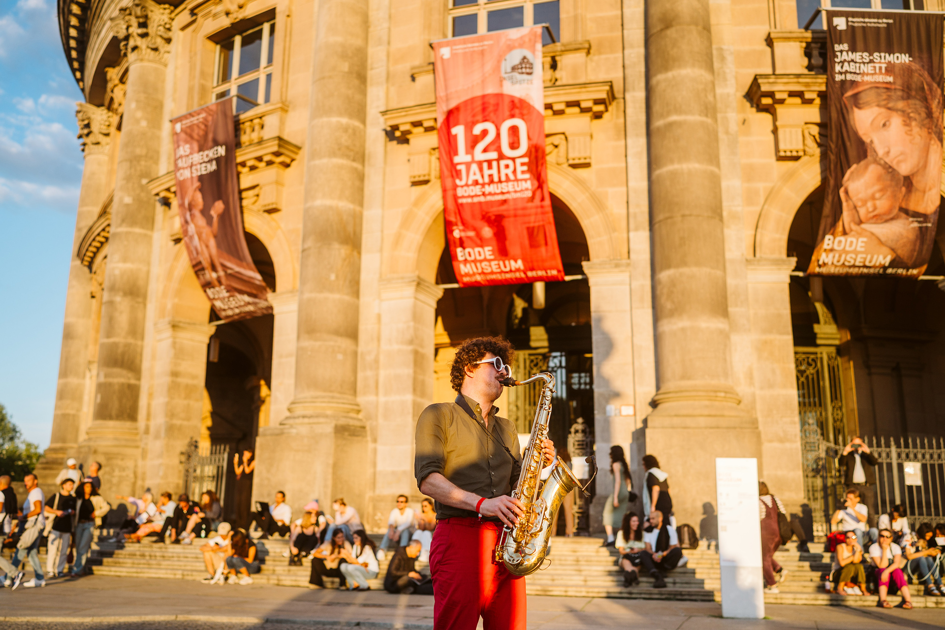A saxophonist plays in front of the Bode Museum, surrounded by many people and colorful posters.