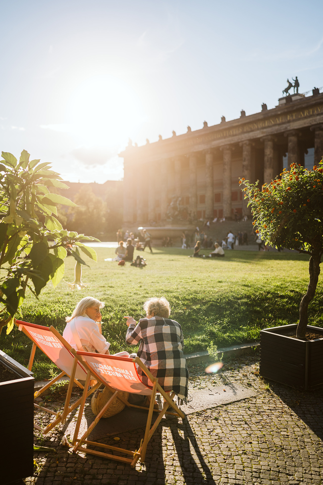 Two women sit in sunlight on lounge chairs in front of a historic building, surrounded by green plants and a lawn.