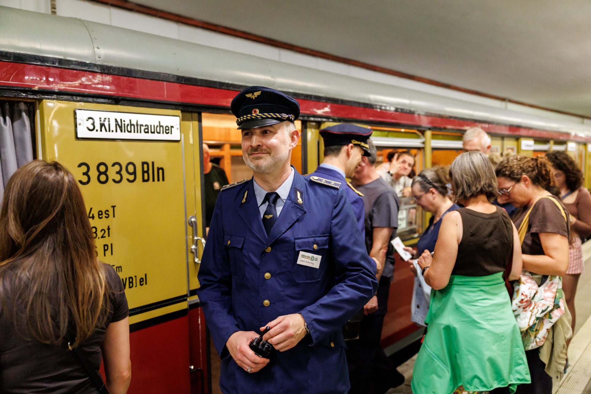 Ein Zugbegleiter steht vor einem alten Zug, während Passagiere am Bahnsteig auf den Zug warten.