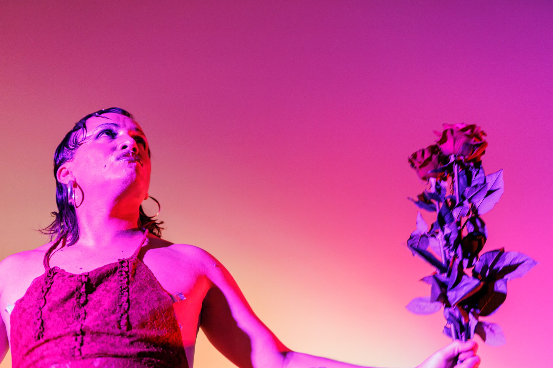Eine Person in buntem Licht hält eine Rose und blickt nach oben, gekleidet in ein schwarzes Kleid.