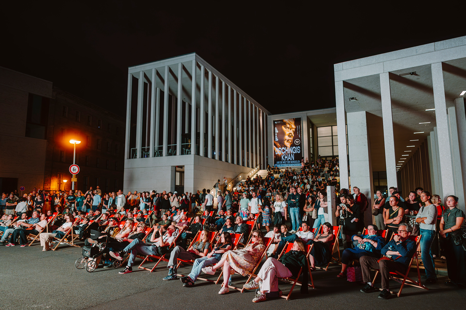 Menschen sitzen in Liegestühlen bei einer nächtlichen Veranstaltung vor einem modernen Gebäude mit Filmplakat.