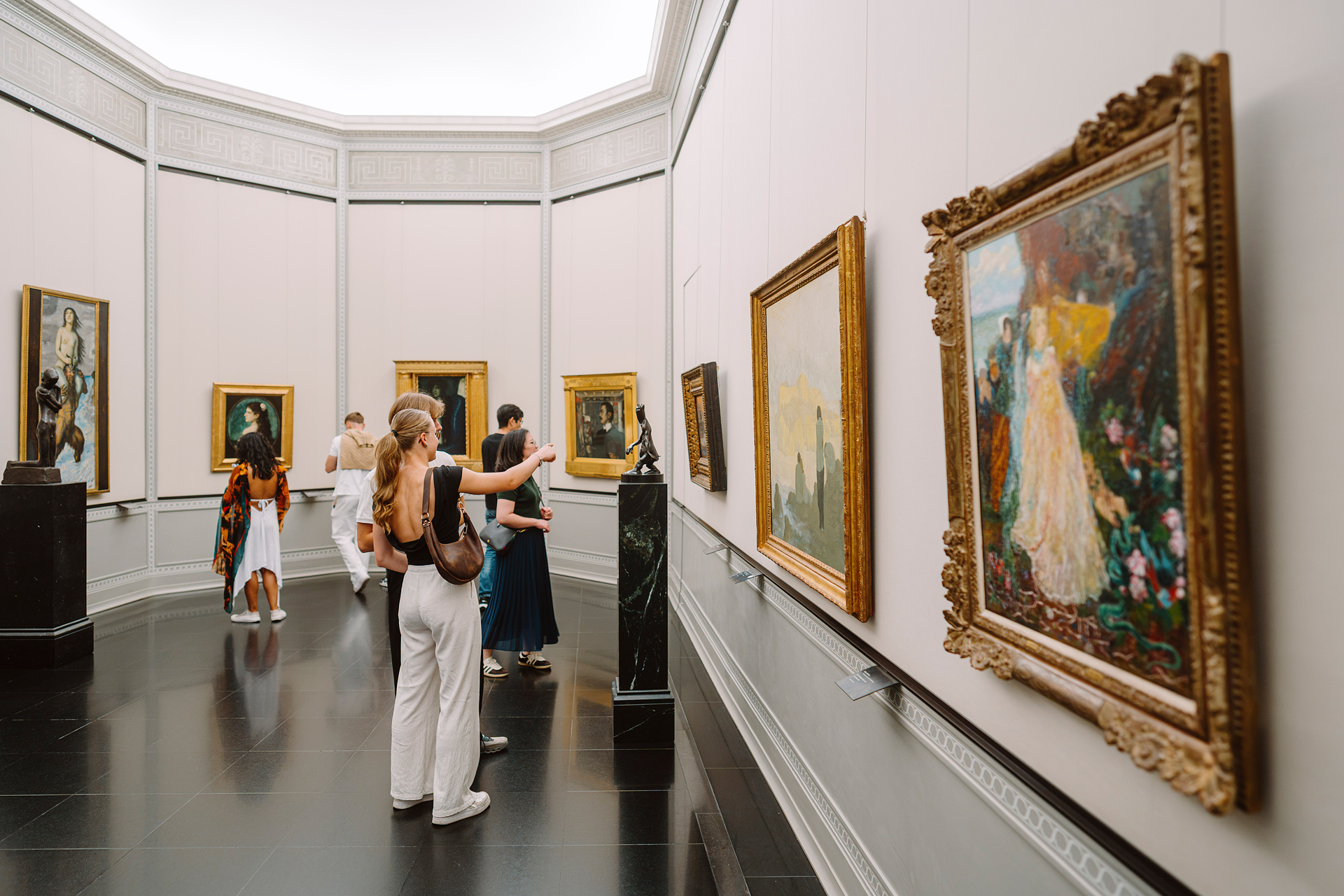 Menschen betrachten Kunstwerke in einem stilvollen Museum mit goldenen Rahmen und modernen Skulpturen.