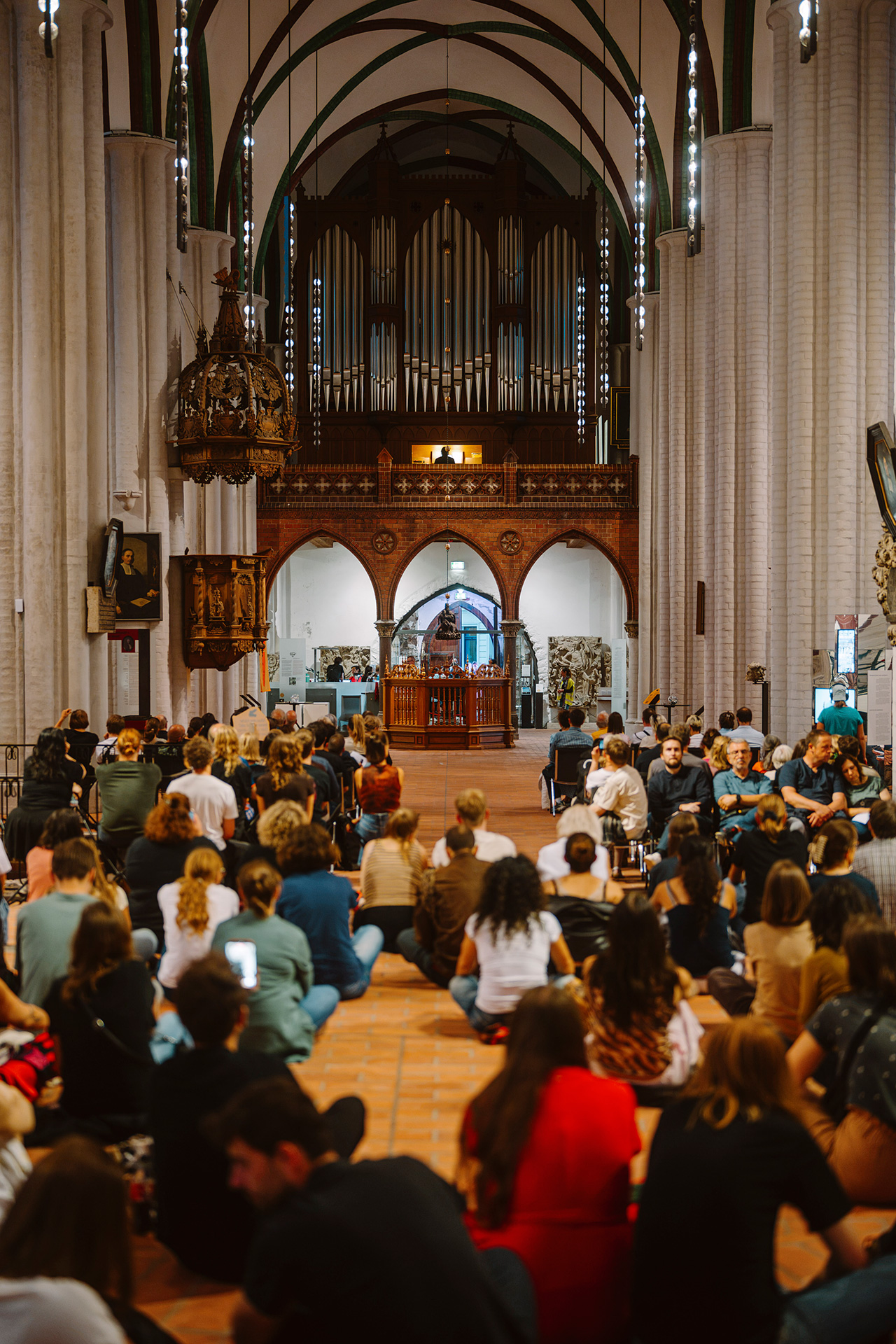 Innere einer Kirche mit versammeltem Publikum, im Hintergrund eine Orgel und kunstvolle Architektur.